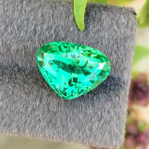 Può includere: Una gemma verde smeraldo, sfaccettata e a forma di cuore, poggia su una superficie grigia e strutturata. Le sfaccettature della gemma riflettono la luce, creando un effetto scintillante. Foglie verdi e fogliame sono visibili sullo sfondo.