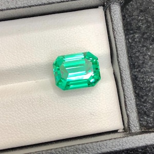 Può includere: Una gemma color verde smeraldo vibrante con un taglio smeraldo rettangolare, caratterizzata da sfaccettature nitide che riflettono la luce. La gemma è posizionata su un inserto bianco imbottito all'interno di una scatola nera.