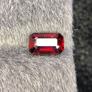 Può includere: Una gemma rosso scuro, tagliata a forma rettangolare, con un interno bianco brillante. La gemma è posta su uno sfondo grigio strutturato, che ne evidenzia il colore vibrante e il taglio.