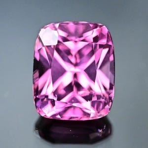 Può includere: Una gemma sfaccettata, taglio a cuscino, in una vibrante tonalità rosa. La superficie della gemma riflette la luce, mostrando la sua intricata struttura interna e gli angoli acuti. La gemma è posta su uno sfondo scuro, evidenziandone la brillantezza.