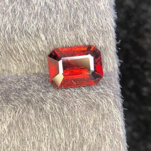 Può includere: Una gemma sfaccettata, taglio rettangolare, di un vibrante colore rosso. La gemma è posta su uno sfondo grigio chiaro, che ne evidenzia i bordi affilati e le sfaccettature interne. Il colore della gemma è uniforme, con una superficie lucida.