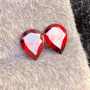 Può includere: Due gemme rosse sfaccettate a forma di goccia con una superficie luminosa e riflettente. Le gemme sono esposte su una superficie morbida e grigia, creando un contrasto tra le trame.