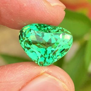 Può includere: Una gemma sfaccettata a forma di cuore, di un vibrante colore verde brillante. La gemma è tenuta tra due dita, mostrando la sua chiarezza e brillantezza. Lo sfondo è sfocato, con fogliame verde visibile.
