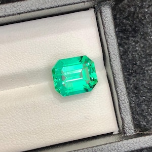Può includere: Una gemma color verde smeraldo brillante con taglio a smeraldo, caratterizzata da una forma rettangolare e superfici sfaccettate. La gemma è esposta all'interno di un piccolo portagioie nero con rivestimento interno bianco.