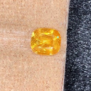 Può includere: Una gemma gialla brillante, taglio a cuscino. La gemma è posta su una superficie strutturata marrone chiaro, con uno sfondo scuro e strutturato. Le sfaccettature della gemma riflettono la luce, creando un aspetto brillante.