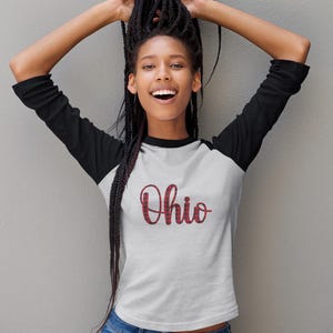 May include: White and black raglan baseball tee with the word "Ohio" in red plaid lettering. The shirt is worn by a person with long black braids and blue jeans.