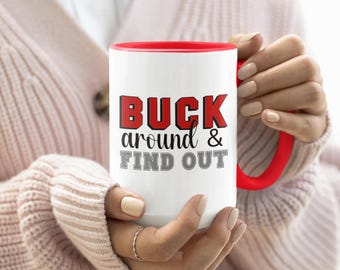 May include: White ceramic mug with a red interior and handle. The mug features the text "BUCK around & FIND OUT" in black and red lettering. The mug is held by a person wearing a pink sweater.