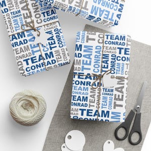 Puede incluir: Varios regalos envueltos con texto "TEAM CONRAD" en azul, gris y blanco. Los regalos están atados con hilo y tienen etiquetas en blanco. También se ven un ovillo de hilo, unas tijeras y etiquetas en blanco.