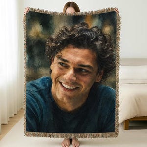 May include: A rectangular woven throw blanket featuring a large, painterly portrait of a smiling person with curly dark hair and a blue shirt. The blanket has a textured, fringed border along all four edges, creating a decorative finish.