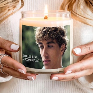 May include: A burning candle in a clear glass jar. The candle has a label with a photo of a young man and the text "Smells Like Second Place". The candle is held by a person wearing a white jumper.