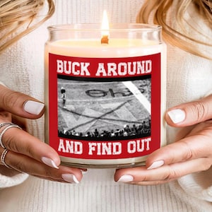 Puede incluir: Una vela de vidrio encendida con una etiqueta roja que dice "BUCK AROUND AND FIND OUT". La etiqueta presenta una imagen en blanco y negro de un campo de fútbol. La vela se sostiene con ambas manos.