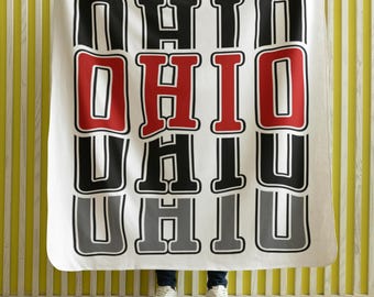 May include: White blanket featuring the word "OHIO" in a vertical, repeating pattern. The letters are black with a grey outline, and the central "OHIO" is red. The blanket is held up by someone wearing white trainers.