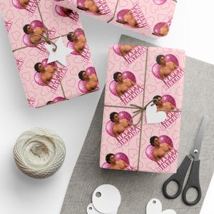 May include: Wrapping paper with a repeating pattern of pink hearts and a shirtless person inside a glittery pink circle, accompanied by the text Happy Birthday. The paper is tied with brown twine and a white star-shaped gift tag.