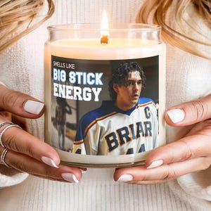 May include: A clear glass jar candle with a lit wick and a label featuring a photograph of a hockey player in a white, blue, and yellow jersey with the text "SMELLS LIKE BIG STICK ENERGY" printed in bold blue and white lettering on the front.