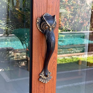 May include: A dark bronze-colored metal door handle shaped like an owl with detailed feathers and large eyes. The handle is mounted vertically onto a door frame with a wood grain finish, featuring a smooth, curved body and a flared tail base.