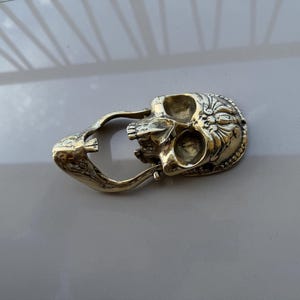 May include: A detailed, gold-colored skull-shaped object with intricate floral and beaded designs. The skull has open eye sockets and a jaw, and is connected to a curved handle. The object is likely a decorative item or a unique accessory.