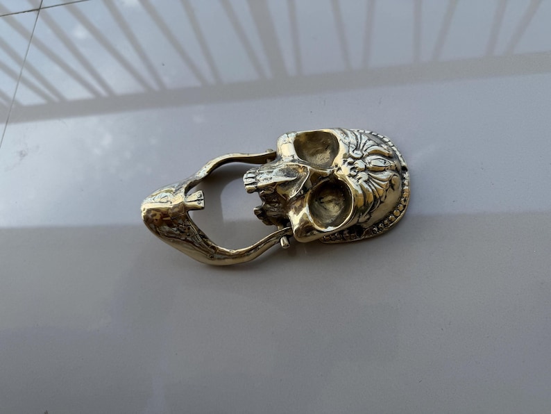 May include: A detailed, gold-colored skull-shaped object with intricate floral and beaded designs. The skull has open eye sockets and a jaw, and is connected to a curved handle. The object is likely a decorative item or a unique accessory.