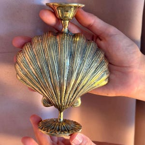 May include: A brass seashell-shaped candle holder. The candle holder has a detailed, textured design with a scalloped shell shape and a circular base. The candle cup is at the top.