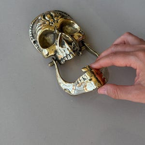 May include: A brass skull-shaped door knocker with intricate details. The knocker features a hinged jaw and a decorative design on the skull's crown. The skull is a golden color.