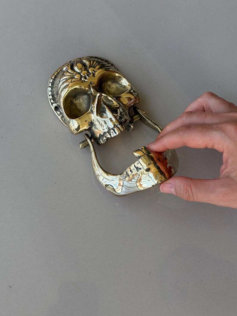 May include: A brass skull-shaped door knocker with intricate details. The knocker features a hinged jaw and a decorative design on the skull's crown. The skull is a golden color.
