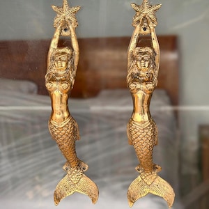 May include: Two golden mermaid door handles. Each handle features a mermaid holding a starfish above her head. The mermaids have detailed scales and flowing hair. The handles are made of a shiny, metallic material.