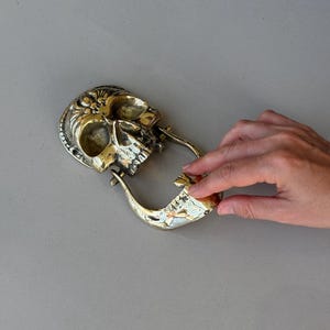May include: A brass skull-shaped belt buckle with intricate detailing. The buckle features a skull design with a hinged jaw and a loop for the belt. The metal has a polished, golden finish, and the design is highly detailed.