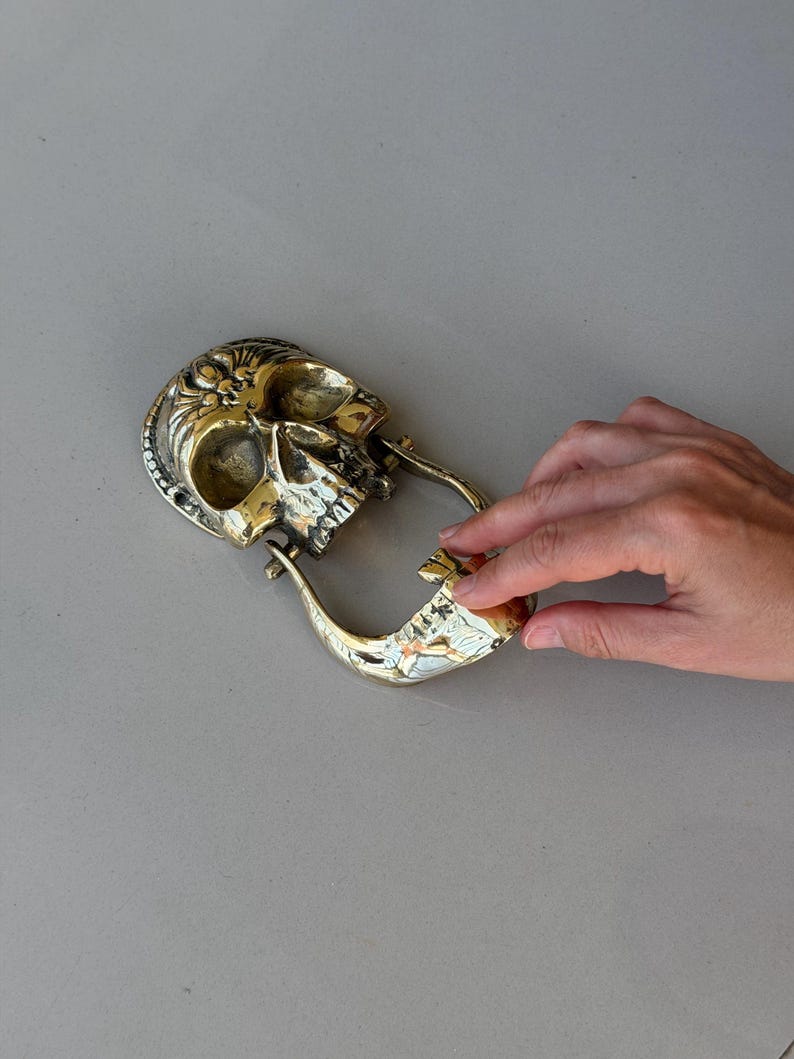May include: A brass skull-shaped belt buckle with intricate detailing. The buckle features a skull design with a hinged jaw and a loop for the belt. The metal has a polished, golden finish, and the design is highly detailed.