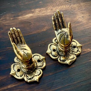 May include: Two brass-colored hand-shaped decorative objects. Each hand is raised in a gesture of blessing and is mounted on a lotus flower base. The objects are made of metal and have a polished finish. The background is a dark wood surface.