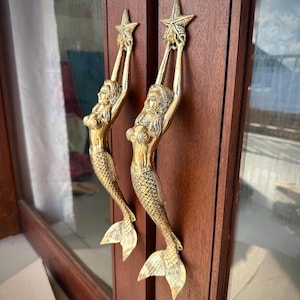 May include: Two brass mermaid door handles. Each handle features a mermaid holding a starfish above her head. The mermaids have detailed scales and flowing hair. The handles are mounted on a dark brown wooden door.