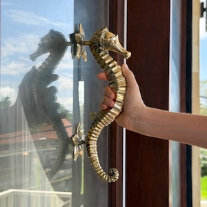 May include: A decorative door handle in the shape of a seahorse, crafted from gold-colored metal. The handle is held by a person, showcasing its intricate design and details. The seahorse handle is mounted on a glass door.