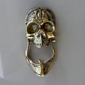 May include: A brass skull-shaped door knocker. The knocker features a detailed skull design with a hinged jaw and a decorative crown. The metal has a polished, golden finish.