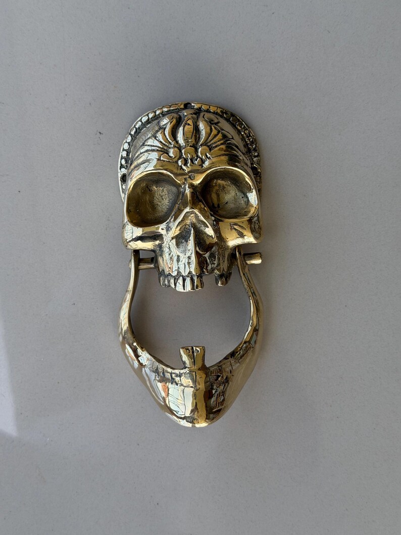 May include: A brass skull-shaped door knocker. The knocker features a detailed skull design with a hinged jaw and a decorative crown. The metal has a polished, golden finish.