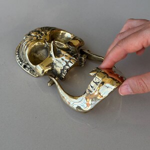 May include: A brass skull-shaped door knocker with intricate details. The knocker features a hinged jaw and a curved handle. The skull has a polished finish, reflecting light.