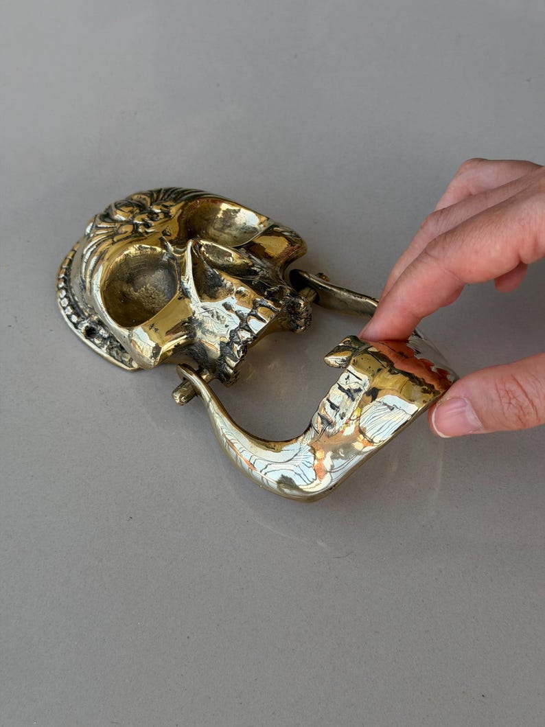 May include: A brass skull-shaped door knocker with intricate details. The knocker features a hinged jaw and a curved handle. The skull has a polished finish, reflecting light.