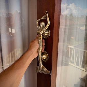 May include: A gold-colored mermaid door handle with a detailed design. The mermaid is depicted with arms raised, and the handle is mounted on a dark wooden door frame. The handle is approximately 12 inches long.