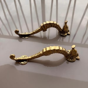 May include: Two brass seahorse-shaped door handles. The handles are gold-colored and feature detailed seahorse designs. The handles are mounted on a light-colored surface, suggesting they are ready for installation on a door or cabinet.