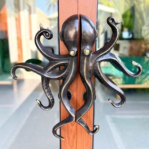May include: A dark bronze-toned metal door handle set shaped like a split octopus. The design features two symmetrical halves, each with four curved tentacles and a central head. The eyes are accented with small, circular, light-colored metallic inlays.
