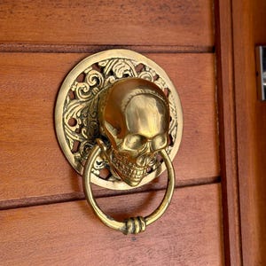 May include: A polished brass door knocker featuring a three-dimensional human skull mounted on a circular backplate with intricate scrollwork detailing. A circular brass ring hangs from the jaw of the skull, providing a functional handle.