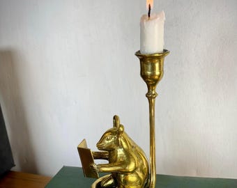 May include: A polished brass candle holder featuring a sculpted mouse sitting and holding an open book. A tall, slender stem rises from the mouse's tail to support a cup holding a single white taper candle with a lit flame, creating a warm glow.