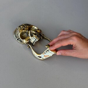 May include: A brass skull-shaped door knocker. The knocker features a detailed skull design with a decorative backplate and a curved handle. The metal has a polished, golden finish, and the knocker is set against a neutral background.
