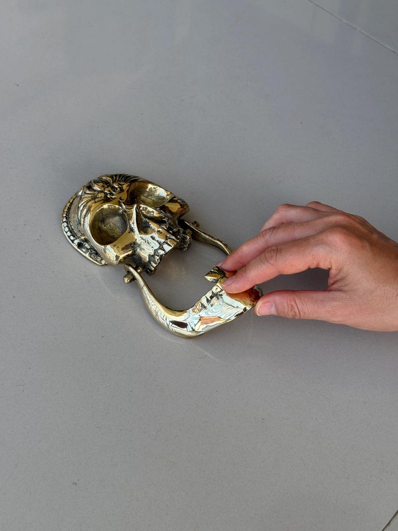 May include: A brass skull-shaped door knocker. The knocker features a detailed skull design with a decorative backplate and a curved handle. The metal has a polished, golden finish, and the knocker is set against a neutral background.
