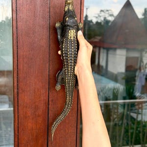 May include: A dark brown and gold-toned metal door handle shaped like a crocodile. The handle features a textured, scaly surface with gold highlights along the back and head, mounted vertically on a dark wooden door frame for functional use.