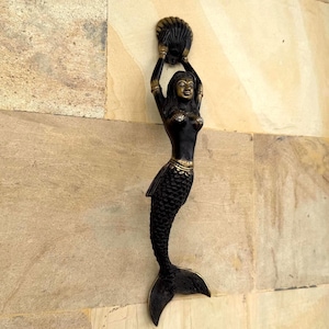 May include: A vertical door handle shaped like a mermaid with a dark bronze finish. The mermaid figure has her arms raised above her head, holding a seashell. Her tail features detailed scales, and she wears a shell-shaped top and arm bands.