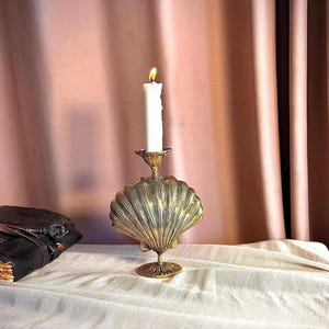 May include: A gold-colored seashell-shaped candle holder with a lit white candle. The candle holder has a detailed, textured design and sits on a round base. The background features a pink curtain and a white tablecloth.
