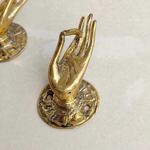 May include: A pair of brass-colored hand-shaped decorative objects. Each hand is in a mudra pose, mounted on a round base with a floral design. The objects are likely used as decorative hardware or wall accents.