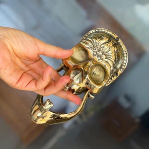 May include: A detailed, golden skull-shaped door knocker. The knocker features intricate floral and beaded designs around the skull's face and a curved handle. The skull's eye sockets and nose cavity are visible. The overall design is ornate and decorative.