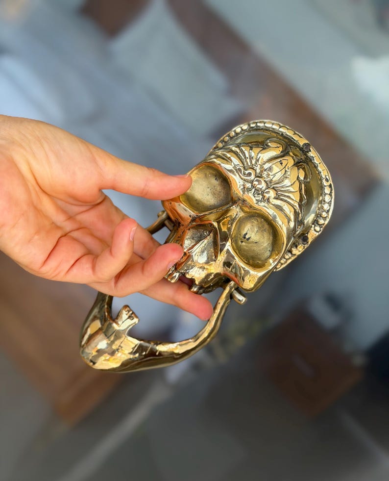 May include: A detailed, golden skull-shaped door knocker. The knocker features intricate floral and beaded designs around the skull's face and a curved handle. The skull's eye sockets and nose cavity are visible. The overall design is ornate and decorative.
