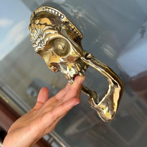 May include: A brass skull-shaped door knocker with intricate details. The knocker features a skull design with a handle. The brass has a polished finish, reflecting light. The knocker is mounted on a glass door.
