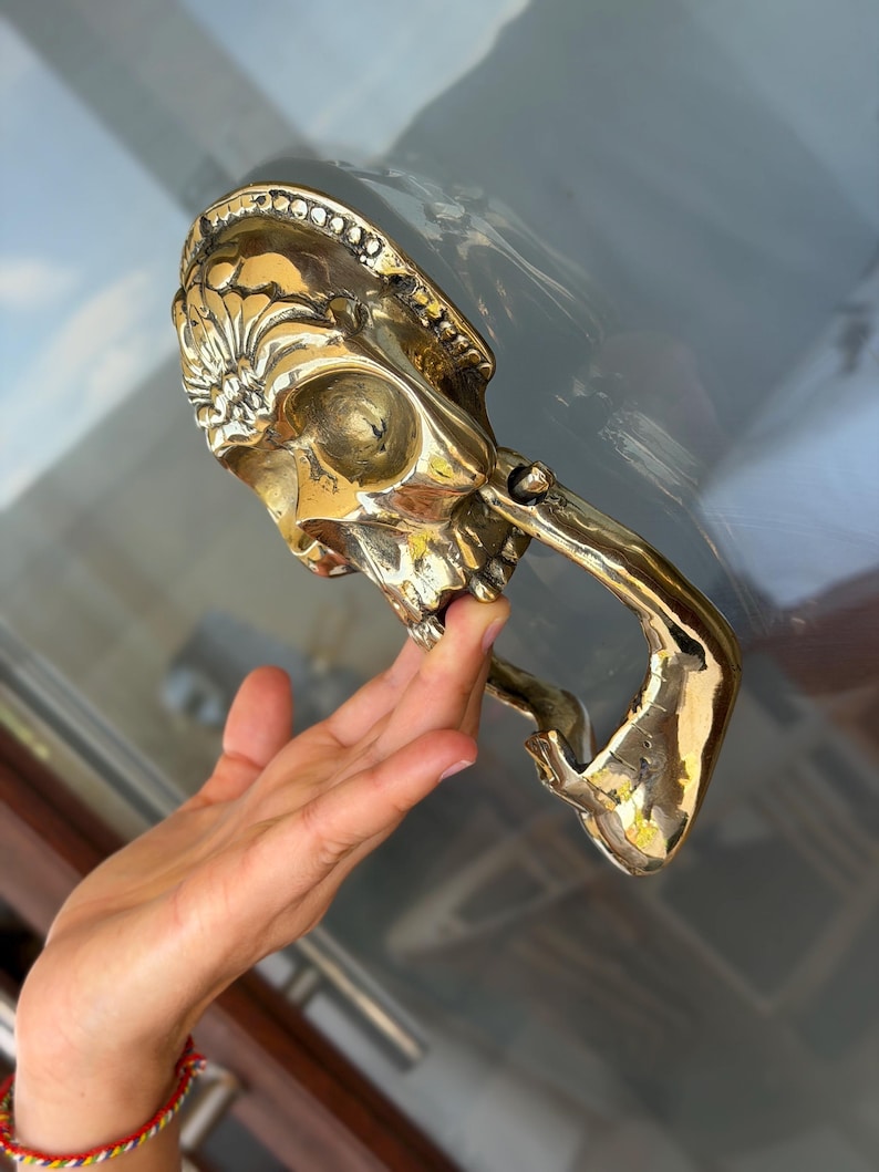 May include: A brass skull-shaped door knocker with intricate details. The knocker features a skull design with a handle. The brass has a polished finish, reflecting light. The knocker is mounted on a glass door.