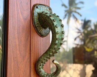 May include: A door handle shaped like an octopus tentacle with a weathered green patina finish. The handle features a textured surface with raised suckers and is mounted on a decorative oval base plate with visible screw holes for installation.